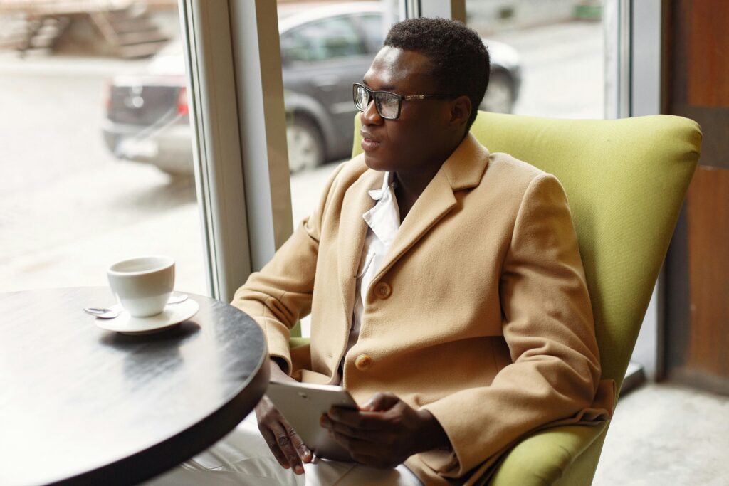 Serious African American male in trendy formal suit and eyeglasses sitting on cozy chair in cafe with cup of coffee and browsing tablet